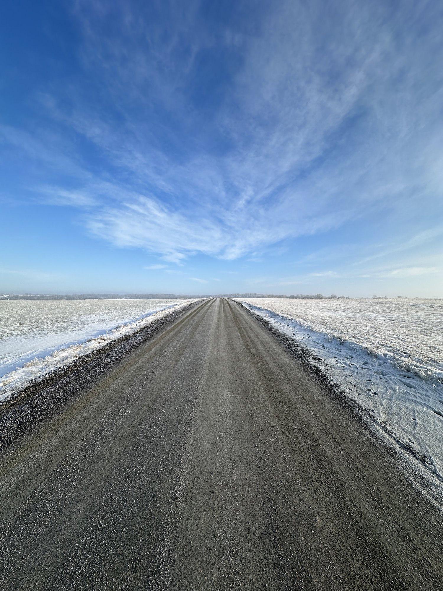 A long, straight road stretches into the horizon flanked by snowy fields under a blue sky.
